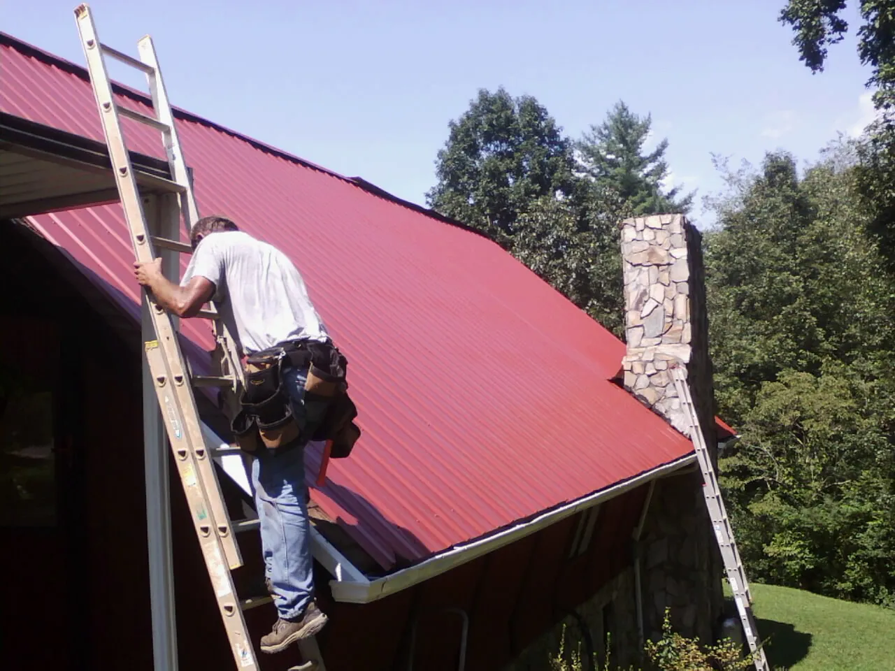 Roofer on a ladder beside a red metal roof and stone chimney — Soffit & Fascia Repair by Roofing Brothers in Newdale